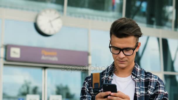 近代的な空港の背景にネットワークでチャット用のスマート フォンを使用してガラスのスタイリッシュな若者。クローズ アップ. — ストック動画