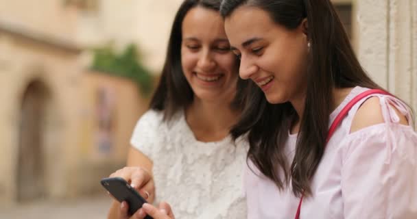 Två leende flickor håller mobilen. De ser glada. Utomhus. — Stockvideo