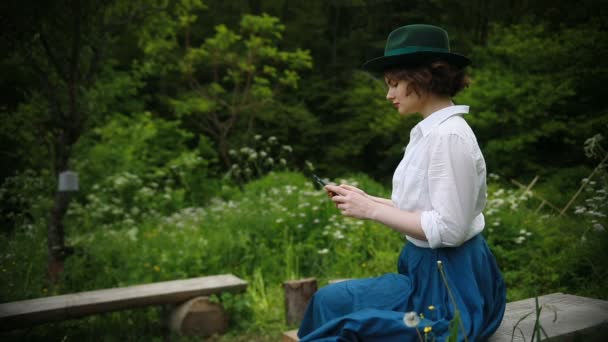 Ung flicka sitter på en bänk i parken med mobiltelefon, använder mobila enheter, foto på bänken i sommaren Park — Stockvideo