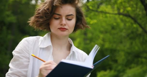 Retrato de cerca de una joven escribiendo en su diario, en el parque. Mujeres en el parque escribe en su diario personal — Vídeos de Stock