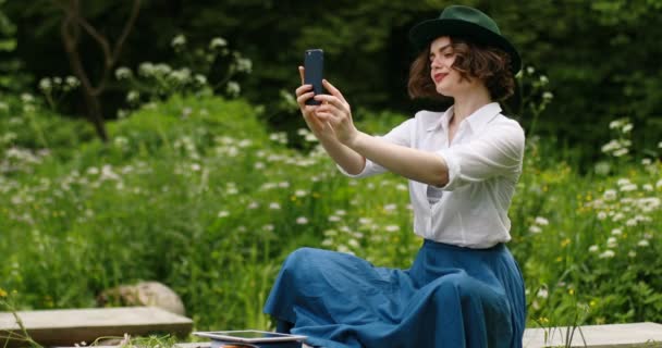 Schöne junge Frauen mit Hut, die auf der Parkbank sitzen und das Smartphone für ein Selfie benutzen — Stockvideo