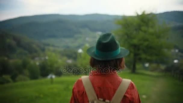Туристична молода жінка в капелюсі ходить по горі. Активна здорова дівчина-хіпстер, що йде в ліс, мандрівник з рюкзаком, що йде по шляху в гори — стокове відео