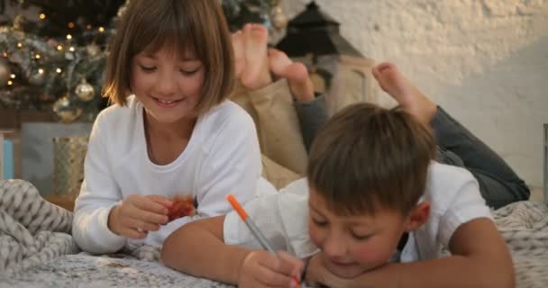 Celebração de festa de Natal, infância. Menino e menina está deitado perto de árvore de natal decorada e escrever carta de Natal para Papai Noel — Vídeo de Stock