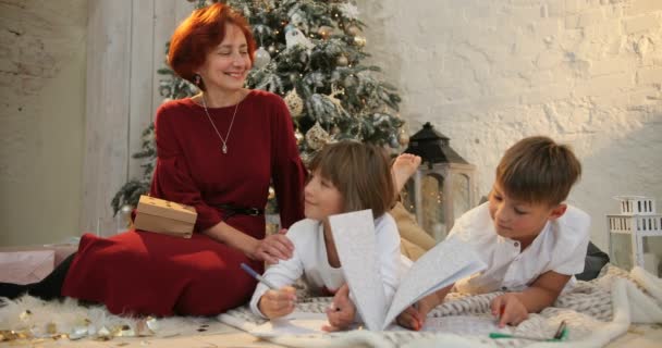 Abuela e hijos cerca del árbol de navidad decorado escribiendo una carta de navidad a Santa Claus. La mañana antes de Navidad. Familia con niños celebrando la Navidad en casa — Vídeo de stock