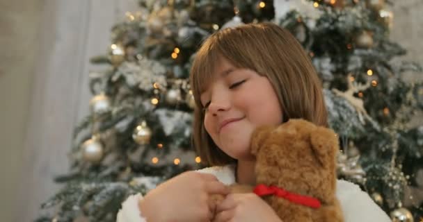 Nahaufnahme Porträt eines lächelnden kleinen Mädchens umarmt Teddybär Weihnachtsgeschenk. Weihnachtsfeier, Kindheit. Weihnachtsbaum-Hintergrund. Weihnachtsurlaub und Neujahr. — Stockvideo