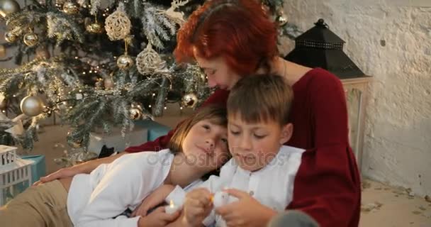 Abuela abrazando a sus nietos cerca del árbol de navidad decorado. La mañana antes de Navidad. Familia con niños celebrando la Navidad en casa — Vídeos de Stock