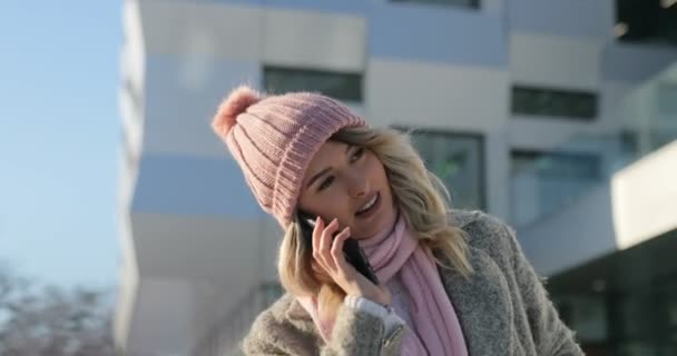 Счастливая девушка разговаривает по телефону, пересекает зимнюю улицу, снежное Рождество. Женщина разговаривает по смартфону в зимнем пальто и розовом шарфе — стоковое видео