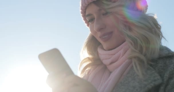 Nära upp skott av flickan med app för smartphone, leende och SMS på mobiltelefon. Kvinnan bär en vinter päls och rosa halsduk och hatt — Stockvideo