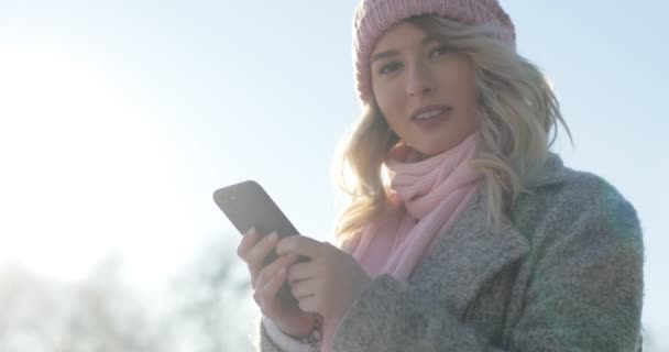 Retrato de niña utilizando la aplicación en el teléfono inteligente, sonriendo y mensajes de texto en el teléfono móvil. Mujer vistiendo un abrigo de invierno y rosa bufanda y sombrero, mirando a la cámara — Vídeos de Stock