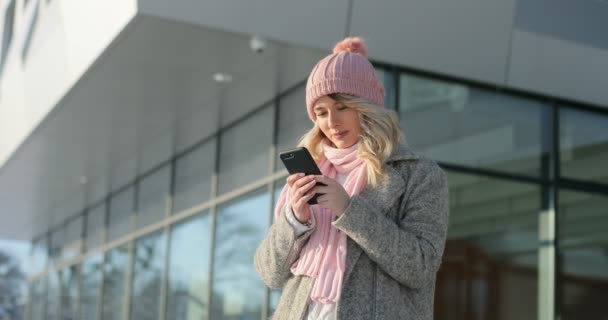 Menina feliz usando aplicativo no smartphone, sorrindo e mensagens de texto no telefone celular. Mulher usando um casaco de inverno e lenço rosa e chapéu — Vídeo de Stock