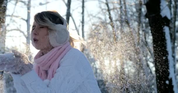 Красива щаслива дівчина дме сніг у морозному зимовому парку. На відкритому повітрі. Літаючі сніжинки, сонячний день. Підсвічування. Радісна красуня розважається в зимовому парку . — стокове відео