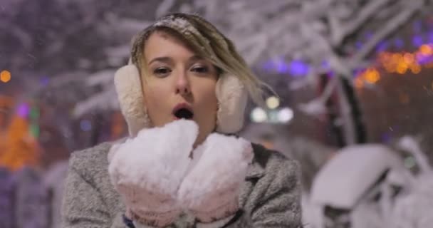 Portrait de belle fille heureuse soufflant de la neige dans la nuit ville d'hiver, femme regardant la caméra. Dame portant des vêtements classiques élégants tricotés d'hiver. . En plein air. Flocons de neige volants , — Video