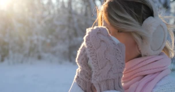 Портрет женщины пьет горячий чай или кофе из чашки в уютном снежном парке зимой. Красивая девушка наслаждается зимой на открытом воздухе с кружкой теплого напитка. Солнечный день, девушка смотрит в камеру — стоковое видео
