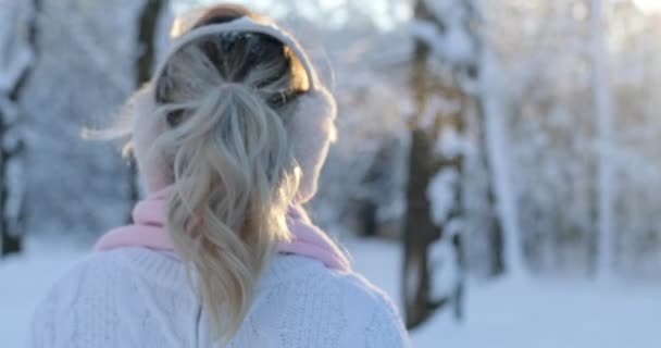 Menina encantadora vestindo um pulôver branco de inverno e cachecol rosa desce o parque de inverno, o sol está brilhando, do que se transforma em câmera e sorri. Mulher desfrutando de neve, expressando positividade — Vídeo de Stock