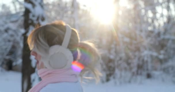 Muchacha atractiva con un suéter blanco de invierno y una bufanda rosa baja por el parque de invierno, el sol está brillando, que se convierte en cámara y sonríe. Mujer disfrutando de las nevadas, expresando positividad — Vídeo de stock