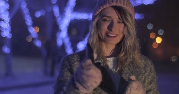Retrato de la joven chica hermosa en la ciudad de invierno de noche. Mujer mostrando teléfono con pantalla, luces de colores, adornos blancos en el fondo, concepto de publicidad con espacio para el texto — Vídeos de Stock
