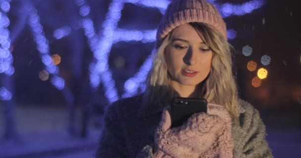 Retrato de la joven chica hermosa en la ciudad de invierno de noche. Mujer mostrando teléfono con pantalla, luces de colores, adornos blancos en el fondo, concepto de publicidad con espacio para el texto — Vídeos de Stock