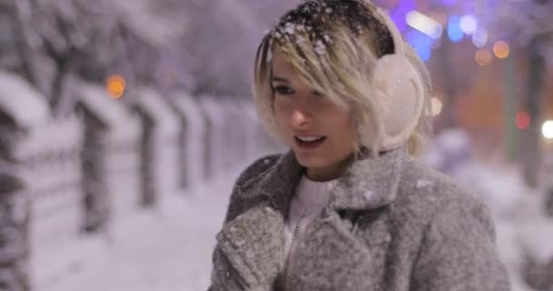 Ritratto di una giovane donna sorridente che indossa dei manicotti per le orecchie, cercando di riscaldarsi nella città invernale notturna. Concetto invernale. Natale, concetto di vacanze invernali. Nevicate . — Video Stock