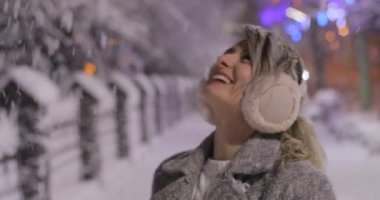 Kendini gece kış şehirde ısıtmak çalışan kulak muffs, giyen bir genç gülümseyen kadın portresi. Kış konsepti. Noel, kış tatil kavramı. Kar yağışı.