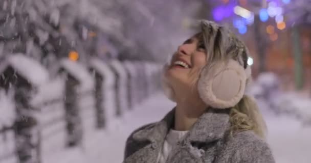 Retrato de uma jovem mulher sorrindo usando abafadores de ouvido, tentando se aquecer na cidade de inverno da noite. Conceito de inverno. Natal, conceito de férias de inverno. Queda de neve . — Vídeo de Stock