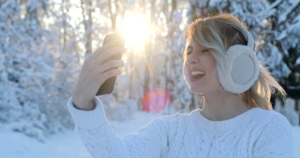 Retrato de una hermosa adolescente con orejeras, tomando una selfie con teléfono inteligente al aire libre en invierno. Navidad, concepto de vacaciones de invierno. Mujer saludando mano a cámara — Vídeos de Stock