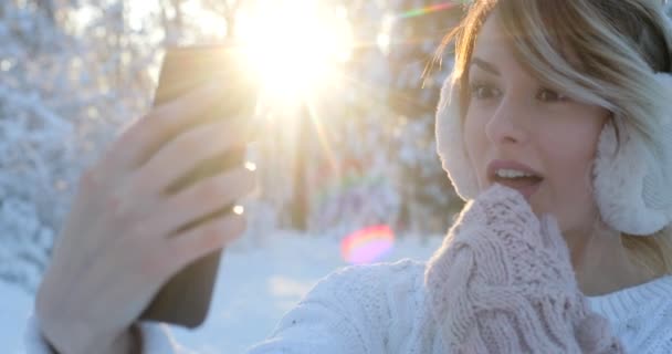 Närbild porträtt av beautifuln tonårsflicka med hörselkåpor, tar en selfie med smartphone utomhus på vintern. Jul, vinter semester koncept. Kvinna viftande hand till kamera — Stockvideo