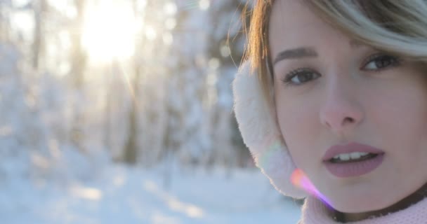 Primer plano retrato de hermosa chica feliz con orejeras, en el parque de invierno helada. Al aire libre. Copos de nieve voladores, día soleado. retroiluminado. Sonriendo a la cámara, alegre humor alegre, emociones — Vídeos de Stock