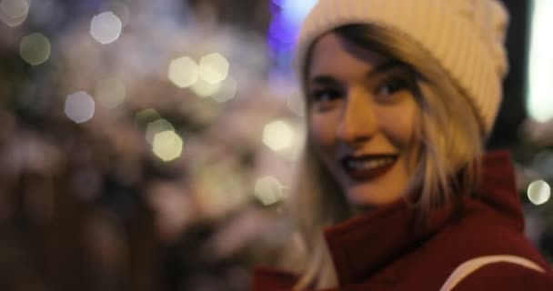 Al aire libre retrato de cerca joven hermosa chica sonriente feliz en la calle de la noche. Feria de Navidad festiva en el fondo. Modelo mirando a la cámara, usando sombrero — Vídeos de Stock