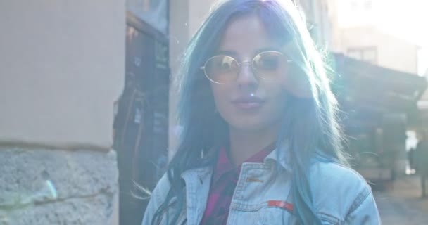 Close up of the young Caucasian girl in hipster style in sunglasses and with blue hair smiling to the camera while wallking a narrow city street in sunlight. Retrato. Fora de casa. — Vídeo de Stock
