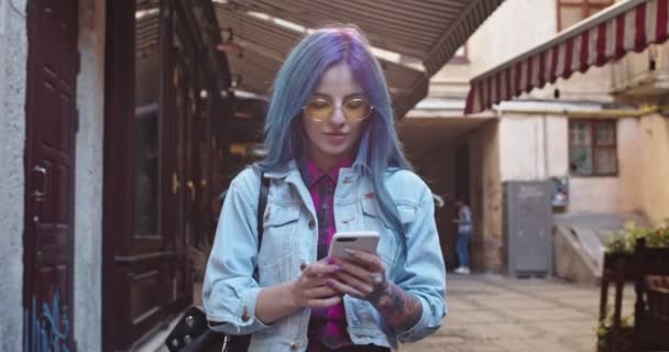 Mladý kavkazský hipster dívka s modrými vlasy procházky po ulici a klepání nebo textování na smartphone jako chatování s někým. — Stock video