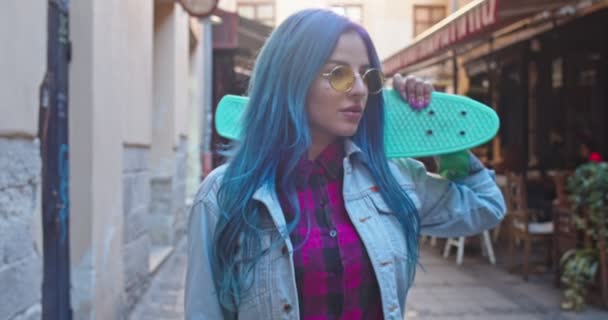 Közelkép a stílusos fiatal csinos hipster lány kék haj tartja gördeszka stnding az utcán a városban, és fordult arc a kamera egy mosollyal. Portré. A kamera ráközelít.. — Stock videók