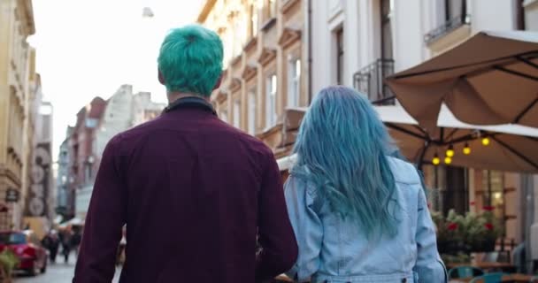 Bakre delen av den unga kaukasiska glada och eleganta par hipsters med blått hår promenader i centrum gatan, sedan vänder sig till kameran och skrattar glatt. Bakifrån. — Stockvideo