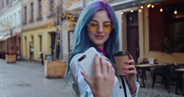 Kavkazské mladé a veselá dívka v hipsterském stylu s modrými vlasy pózovat na smartphone kamery, zatímco pořízení selfie fotky s šálkem kávy jít na ulici ve městě. Zavřít. Venku. — Stock video