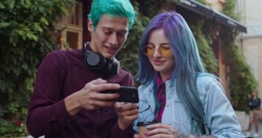 İkisi de sokakta dikilirken, beyaz tenli, mavi saçlı genç kız arkadaşına akıllı telefonuyla bir şey gösteriyor..