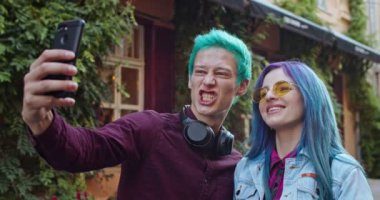 Mutlu ve neşeli genç bir çift mavi saçlı beyaz hippi dışarıda fotoğraf çekerken akıllı telefon kamerasına komik ve alaycı yüzler çiziyor..