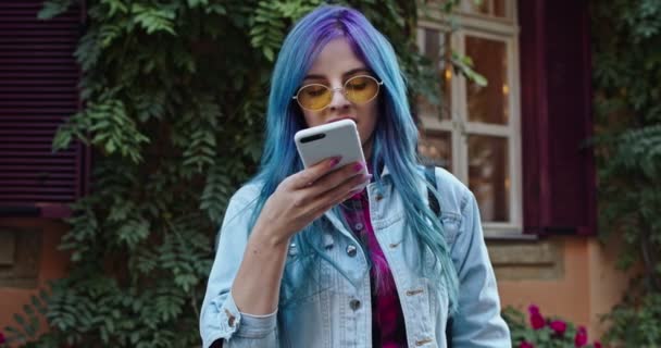 青い髪の若い白人のヒップスタースタイリッシュな女の子は、彼女のスマートフォンに音声メッセージを記録し、素敵な通りでそれを送信します。屋外. — ストック動画