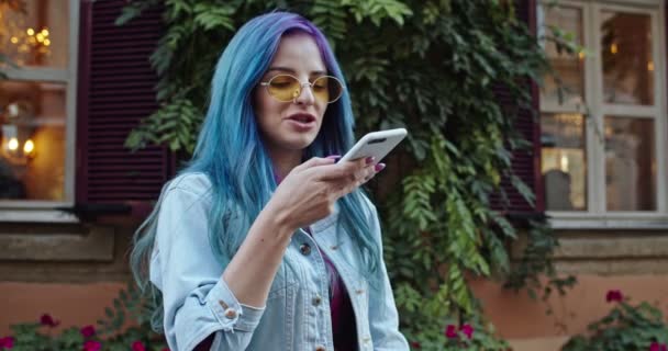 Bella allegra ragazza caucasica giovane hipster in stile ragazza con i capelli blu e in occhiali da sole parlare e registrare un messaggio vocale al telefono. Invio messaggio vocale. Fuori. . — Video Stock