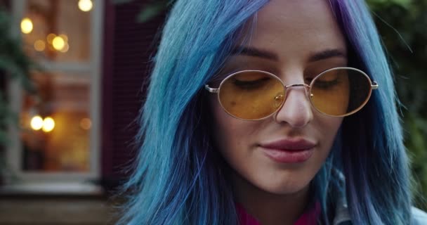 Close up of the face of the young pretty Caucasian hipster girl with blue hair and in sunglasses looking straight to the camera and smiling happily. Outdoor. Portrait. — 비디오