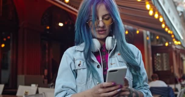 Caucasiana muito jovem hipster menina com cabelo azul e em fones de ouvido tocando e rolando no smartphone, mensagens de texto, em seguida, sorrindo para a câmera. Fora . — Vídeo de Stock