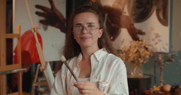 Portrait de la belle blanche heureuse jeune artiste en lunettes souriant joyeusement à la caméra avec un pinceau à la main dans son atelier . — Video