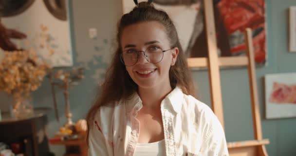 Portrait de la charmante jeune femme artiste caucasienne joyeuse en lunettes souriante et regardant droit vers la caméra dans son atelier. Gros plan . — Video