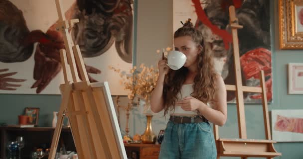 Young caucasian pretty woman artist sitting at the easel and looking at her painting while resting and drinking hot tea or coffee in her workshop. — ストック動画