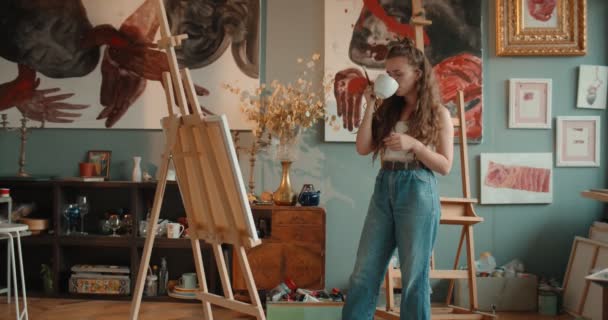 Caucasian attractive young female painter standing at the easel and looking at her piicture while having a rest and sipping hot tea or coffee in her art studio. — ストック動画