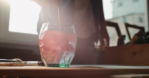 Gros plan du verre d'eau sur la table à la lumière du soleil d'une fenêtre et le peintre masculin laver son pinceau de peinture rouge en elle . — Video