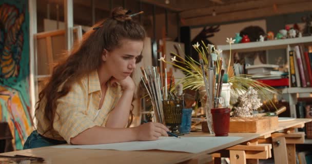 Young Caucasian creative and stylish female artist drawing a sketch for the future painting while working in the workshop. — Stock Video