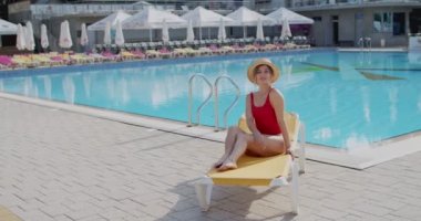 Şapkalı ve kırmızı mayolu güzel beyaz kadın tatilde yüzme havuzunda iskemlede oturuyor..