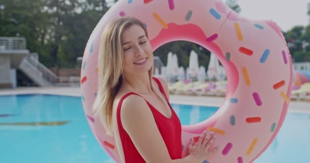Retrato de la joven caucásica guapa con el pelo rubio y traje de baño rojo sonriendo a la cámara y sosteniendo una carroza rosa en la piscina en el verano. Exterior . — Vídeo de stock
