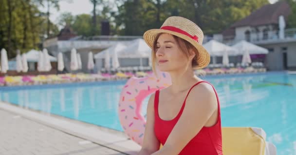 Portraitaufnahme der jungen kaukasischen hübschen und glücklichen Frau mit Hut und rotem Badeanzug, die es sich im Freien am Pool gemütlich macht und in die Kamera lächelt. — Stockvideo
