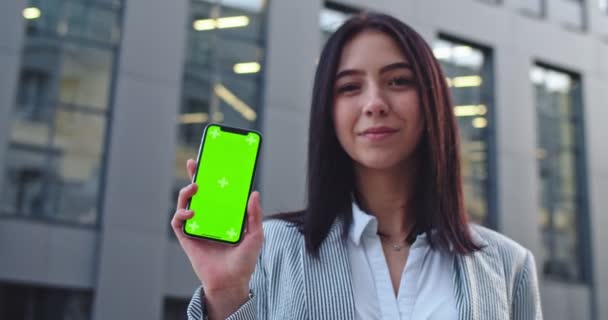 Retrato de la hermosa mujer joven caucásica sonriendo a la cámara y demostrando teléfono inteligente con pantalla verde y movimiento de seguimiento. Clave de croma. Al aire libre . — Vídeos de Stock