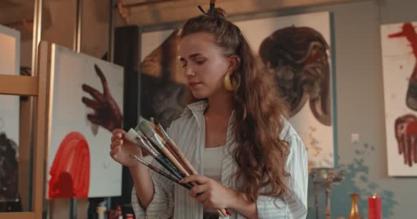 Retrato tiro da jovem pintora caucasiana atraente segurando pincéis em sua mão e olhando para eles, em seguida, sorrindo alegremente para a câmera no estúdio de arte. — Vídeo de Stock